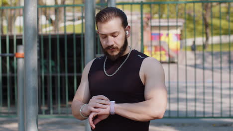 Athletic-lebanese-man-in-sportswear-using-smart-watch,-listening-music-on-earphones-on-playground