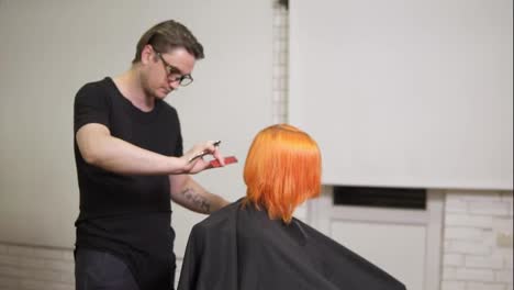 Stylish-professional-male-hairdresser-in-glasses-combing-hair-of-female-client-in-hair-salon-turning-around-her-chair-and