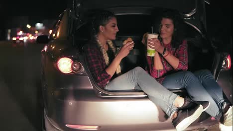 Handsome-girlfriends-sitting-in-the-open-trunk-of-the-car,-talking-and-taking-sandwiches,-drinking-from-the-paper-cup-during-the