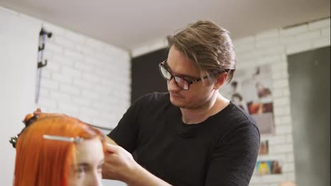 Stylish-male-hairdresser-in-glasses-is-cutting-redhead-woman's-hair-in-beauty-salon.-Young-woman-getting-her-haircut-and