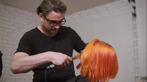 Male-professional-hairdresser-is-straightening-bright-red-woman's-hair-using-a-hairstraightener-in-hair-salon.-Slow-Motion-shot