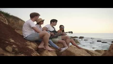 Group-of-young-hipster-friends-sitting-on-the-rocks-by-the-seashore-and-playing-guitar-and-singing-songs.-Slow-Motion-shot
