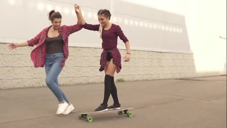 Laughing-young-hipster-girl-being-taught-skateboarding-by-a-friend-who-is-supporting-her-holding-her-hand.-Slow-Motion-shot