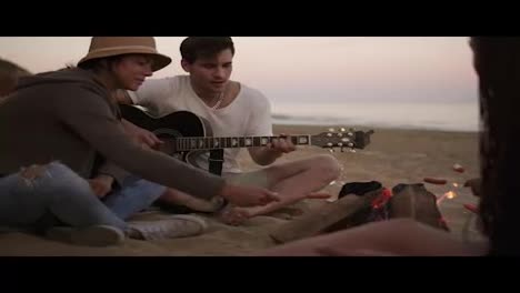 Young-man-is-playing-guitar-by-the-fire-sitting-on-the-beach-together-with-friends.-His-girlfriend-grilling-a-sausage-for-him
