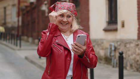 Ältere-Frau-Nutzt-Ihr-Mobiles-Smartphone-Und-Feiert-Den-Gewinn-Einer-Guten-Nachrichtennachricht-Im-Freien-In-Der-Stadtstraße