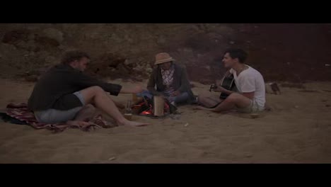 Young-friends-sitting-by-the-fire-on-the-beach-in-the-evening,-grilling-sausages,-drinking-beer-and-playing-guitar.-Slow-Motion