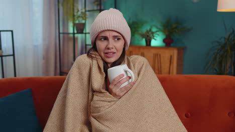 Sick-young-woman-wear-hat-wrapped-in-plaid-sit-alone-shivering-from-cold-on-sofa-drinking-hot-tea