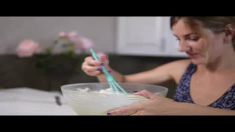 Young-woman-prepares-dough-mixing-ingredients-in-the-the-bowl-using-whisk-in-the-kitchen-and-looking-in-the-camera.-Homemade