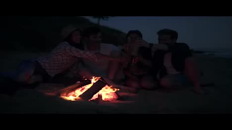 Picnic-of-young-people-with-bonfire-on-the-beach-in-the-evening-drinking-beer-and-doing-cheers.-Happy-friends-singing-songs-and