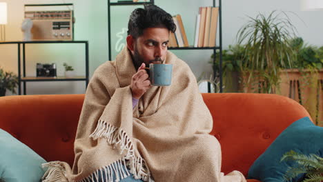 Sick-indian-man-wrapped-in-plaid-shivering-from-cold-drinking-hot-tea-in-unheated-apartment-due-debt