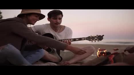 Group-of-young-friends-sitting-by-the-fire-on-the-beach,-grilling-sausages-and-playing-guitar.-Slow-Motion-shot