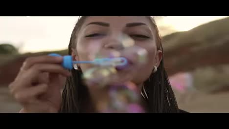 Young-female-hipster-with-dreads-cheerfully-making-soap-bubbles-on-the-beach-in-the-evening.-Slow-Motion-shot