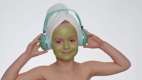 Smiling-young-child-girl-moisturizing-green-mask-on-face-listening-to-music-on-headphones,-dancing