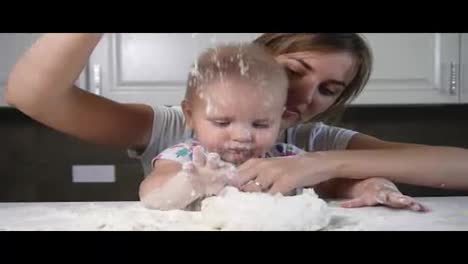 Young-mother-and-her-little-child-preparing-dough-and-pouring-flour-on-the-table.-Little-baby-playing-with-flour.-Family-girls