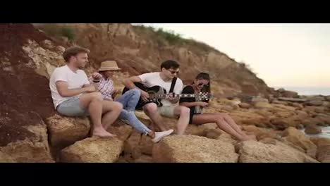 Group-of-young-hipster-friends-sitting-on-the-rocks-by-the-seashore-and-playing-guitar,-singing-songs-and-dancing.-Slow-Motion