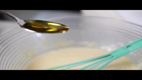Close-Up-view-of-female-hands-adding-oil-from-the-spoon-to-the-bowl-preparing-dough-and-mixing-the-ingredients-using-whisk