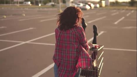 Back-view-of-a-young-woman-sitting-in-the-grocery-cart,-while-her-friend-is-pushing-her-behind-in-the-parking-by-the-shopping