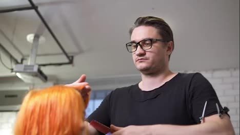 Professional-stylish-hairdresser-in-glasses-is-straightening-bright-red-woman's-hair-using-a-hairstraightener-in-hair-salon