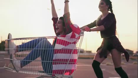 Young-hipster-teen-girls-having-fun-at-the-shopping-mall-parking,-riding-in-shopping-cart-holding-the-american-flag.-Lens-flare