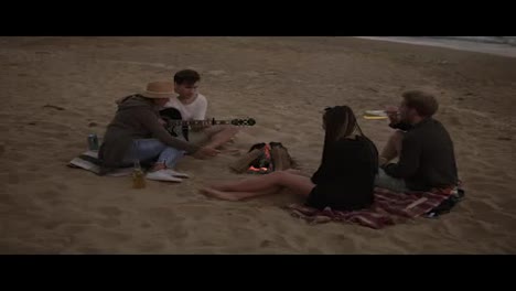 Young-friends-sitting-by-the-fire-on-the-beach,-grilling-sausages,-drinking-beer-and-playing-guitar.-Slow-Motion-shot