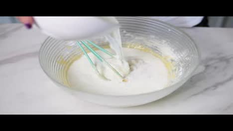 Close-Up-view-of-female-hands-pouring-milk-to-the-bowl-preparing-dough-and-mixing-the-ingredients-using-whisk.-Homemade-food