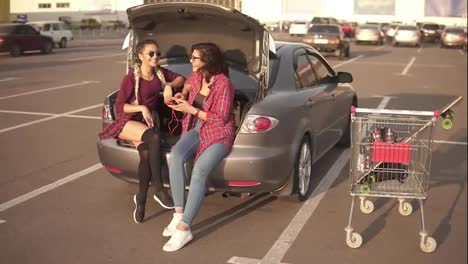 Two-young-attractive-women-in-sunglasses-sitting-inside-of-the-open-car-trunk-and-listening-to-the-music-in-the-smartphone