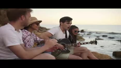 Group-of-young-hipster-friends-sitting-on-the-rocks-by-the-seashore-and-playing-guitar-and-singing-songs.-Slow-Motion-shot