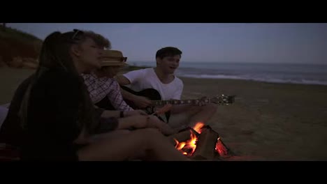 Gruppe-Junger-Und-Fröhlicher-Menschen,-Die-Abends-Am-Feuer-Am-Strand-Sitzen,-Würstchen-Grillen-Und-Gitarre-Spielen