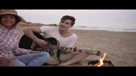 Young-man-is-playing-guitar-by-the-fire-sitting-on-the-beach-together-with-friends.-His-girlfriend-bringing-a-grilled-sausage