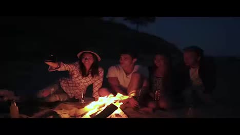 Picnic-of-young-people-with-bonfire-on-the-beach-in-the-evening.-Cheerful-friends-taking-pictures-on-the-phone.-Slow-Motion-shot