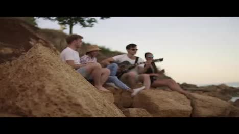 Group-of-young-cheerful-people-sitting-on-the-rocks-by-the-seashore-and-playing-guitar,-singing-songs-and-dancing.-Slow-Motion