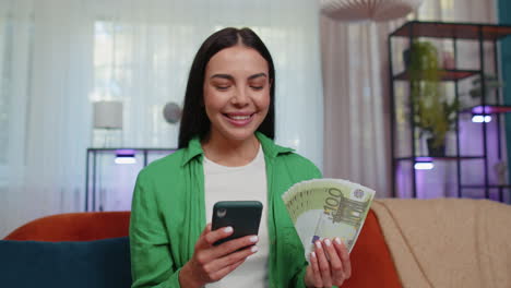 Smiling-happy-young-woman-at-home-counting-money-cash-use-smartphone,-income,-saves,-lottery-win