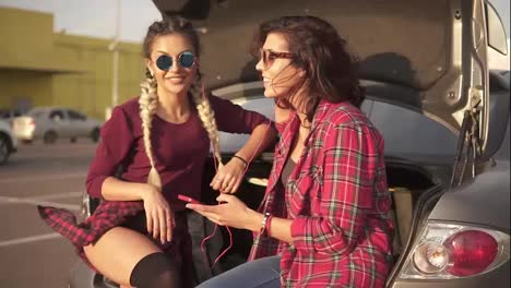 Two-young-attractive-women-in-sunglasses-sitting-inside-of-the-open-car-trunk-in-the-parking-by-the-shopping-mall-and-listening