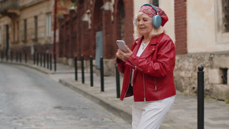 Happy-senior-woman-in-wireless-headphones-choosing,-listening-music-in-smartphone-dancing-outdoors