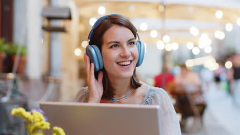 Happy-young-woman-in-wireless-headphones-choosing,-listening-music-dancing-outdoors-city-restaurant