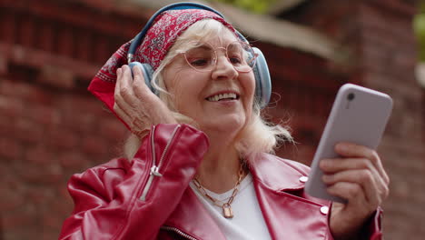 Happy-senior-woman-in-wireless-headphones-choosing,-listening-music-in-smartphone-dancing-outdoors