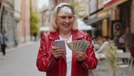 Rich-elderly-woman-counting-money-dollar-cash,-use-smartphone-calculator-app-in-urban-city-street