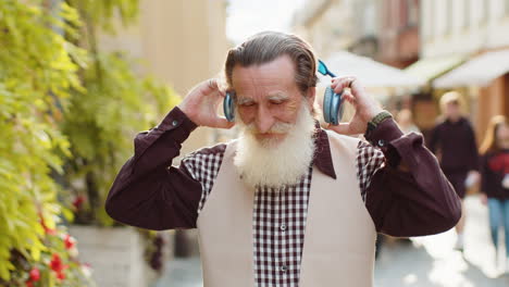 Happy-elderly-old-man-in-wireless-headphones-choosing,-listening-music-dancing-outdoors-city-street