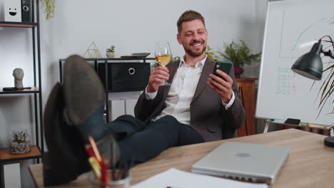 Businessman-drinking-champagne-from-glass-after-working,-putting-legs-on-table-sitting-at-office