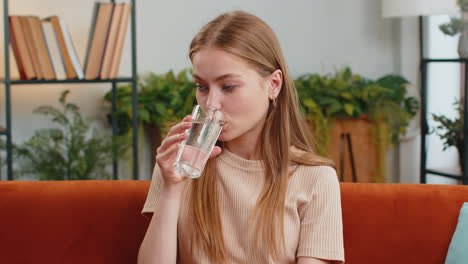 Thirsty-young-woman-sitting-at-home-holding-glass-of-natural-aqua-make-sips-drinking-still-water