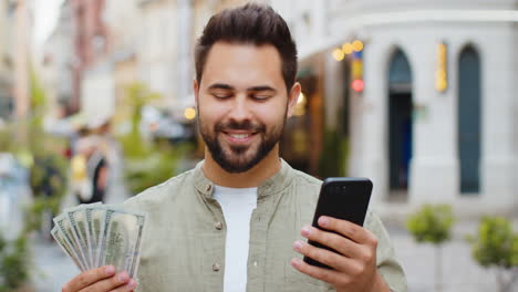 Happy-young-man-counting-money-dollar-cash,-use-smartphone-calculator-app-in-urban-city-street