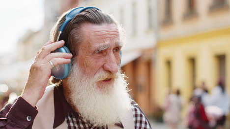 Happy-senior-old-man-in-wireless-headphones-choosing,-listening-music-dancing-outdoors-city-street