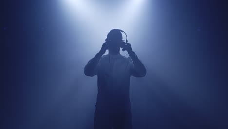 Backlit-silhouette-of-dj-man-standing-in-dark-nightclub-disco,-putting-headphones-on-to-listen-music