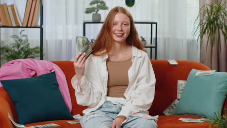 Happy-rich-teenager-child-girl-sits-on-couch-full-of-dollar-cashmoney,-high-profits-lottery-game-win