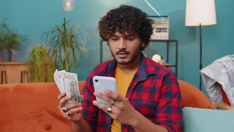 Smiling-happy-indian-man-counting-money-cash-use-smartphone,-income-saves-lottery-win-budget-at-home