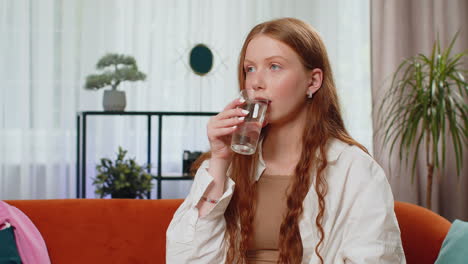 Thirsty-one-teen-girl-sitting-at-home-holding-glass-of-natural-aqua-make-sips-drinking-still-water