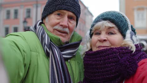 Couple-of-senior-tourists-taking-selfie-in-winter-city-center-smiling,-looking-at-camera,-blog,-vlog