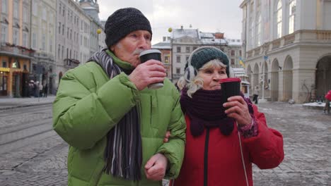 Senior-old-tourists-grandmother-grandfather-walking,-drinking-hot-drink-mulled-wine-in-city-center