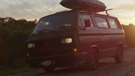 A-surfer-pulls-up-in-a-camper-van-with-a-board-on-top-at-daybreak-on-a-dirt-road-in-a-coastal-area