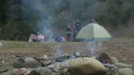 Ein-Erloschenes-Lagerfeuer-Glimmt,-Während-Sich-Camper-Im-Hintergrund-In-Der-Nähe-Eines-Zeltes-Bewegen